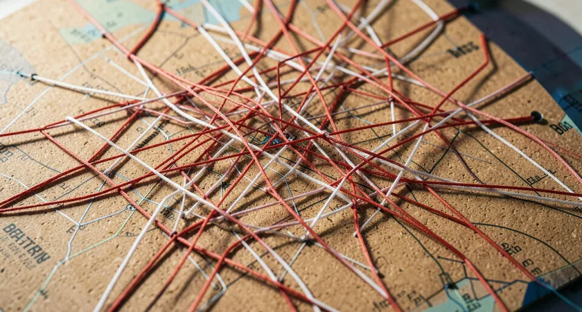 A close-up, top-down view of a dense, tangled network of red and white strings pinned to a corkboard map of a city. The strings connect various districts, illustrating complexity and potential for misconnection, with soft daylight casting sharp shadows.